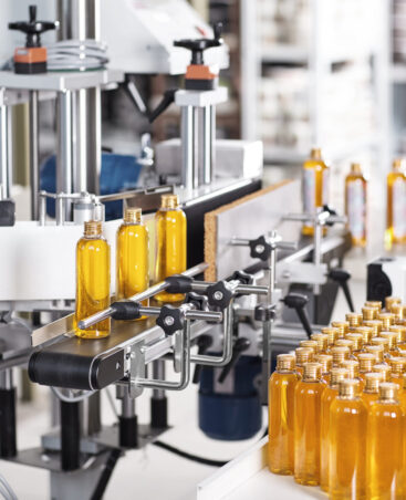 Horizontal shot of cosmetics or pharmacy plant with automated equipment. Transparent plastic bottles filled with yellow substance standing on desk and conveyor line, ready for transportation Horizontal shot of cosmetics or pharmacy plant with automated equipment. Transparent plastic bottles filled with yellow substance standing on desk and conveyor line, ready for transportation